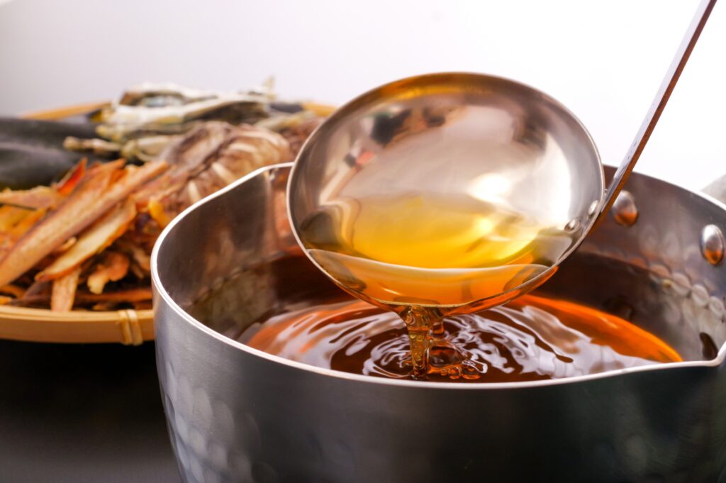 Golden Japanese dashi broth being scooped from a pot, with kombu and dried fish used for making the soup base
