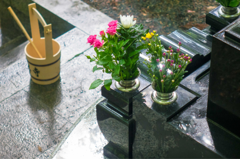 A Japanese gravesite with fresh flowers and a wooden water bucket, reflecting the tradition of visiting ancestors during Ohigan.