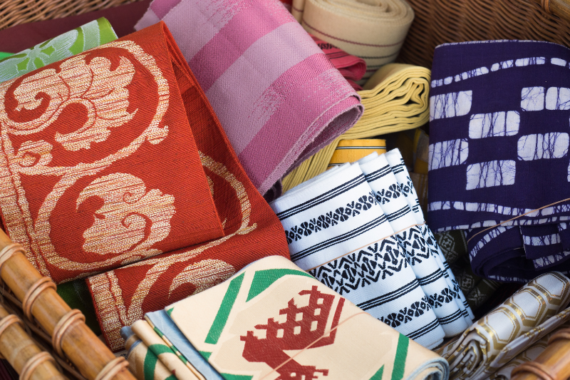 Multiple obi in red, purple, pink, white, and green arranged in a basket, showing different textures and designs.