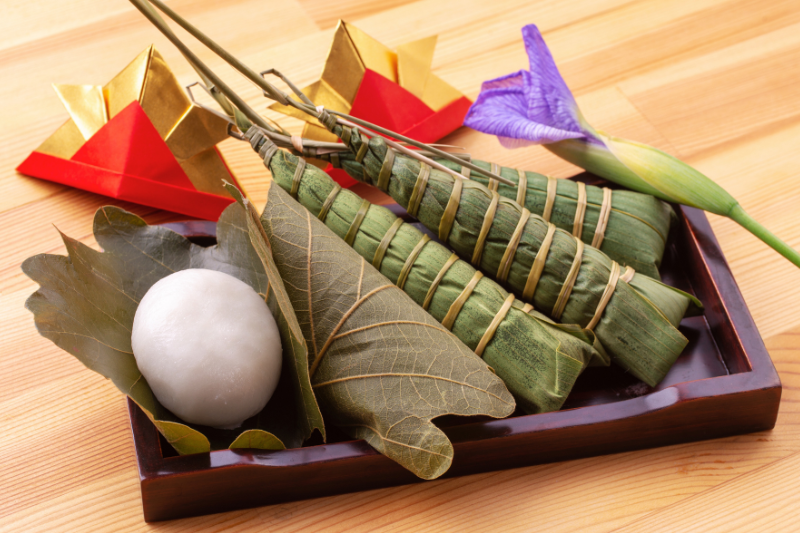 A tray with kashiwa-mochi and chimaki, along with origami samurai helmets and an iris flower, showing traditional foods and decorations for Children’s Day in Japan.