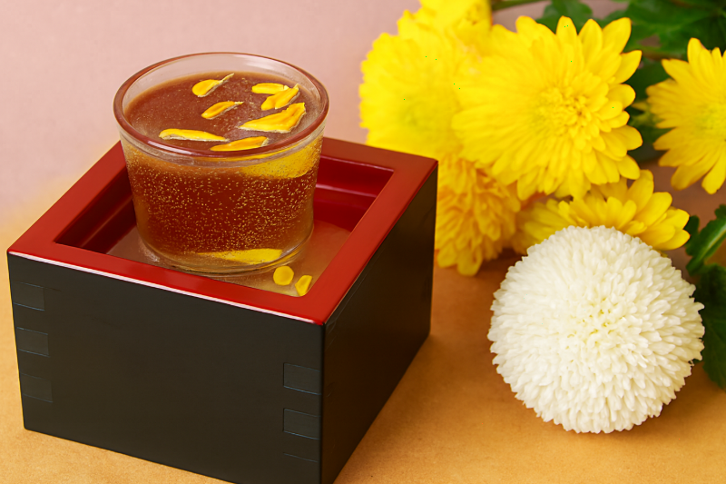 A glass of chrysanthemum sake with floating petals, placed in a wooden masu with yellow and white chrysanthemums in the background.