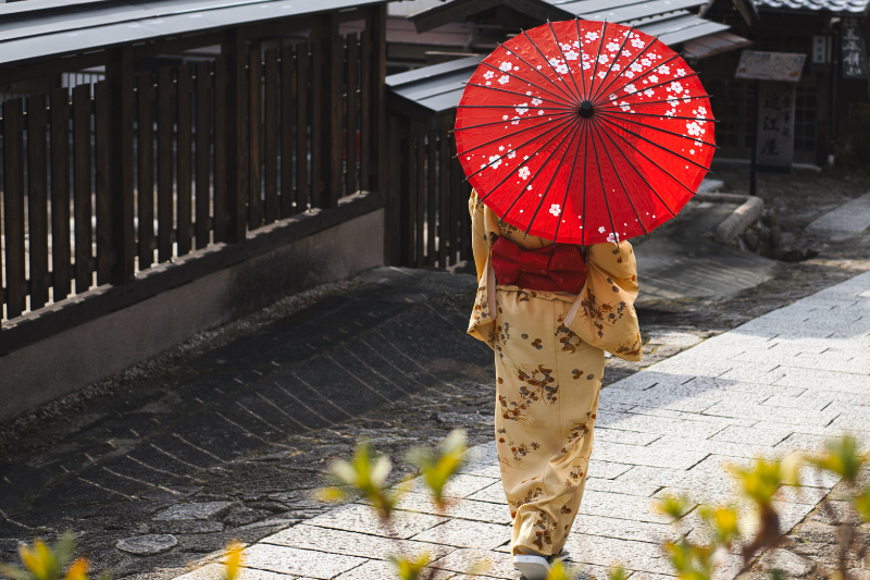 Person in a floral kimono holding a red parasol, walking through a traditional Japanese street.