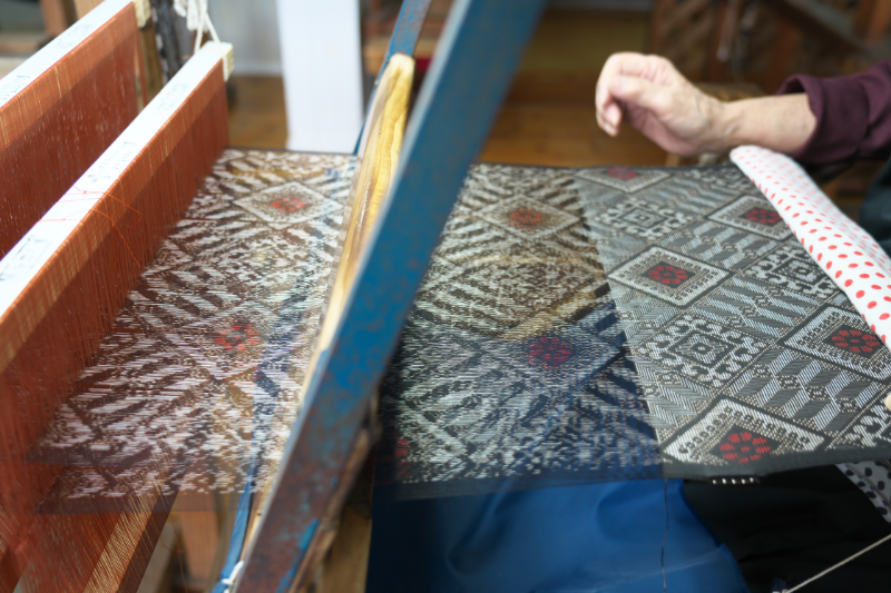 A weaver operating a handloom, creating a textile with red, white, and black geometric and floral patterns.