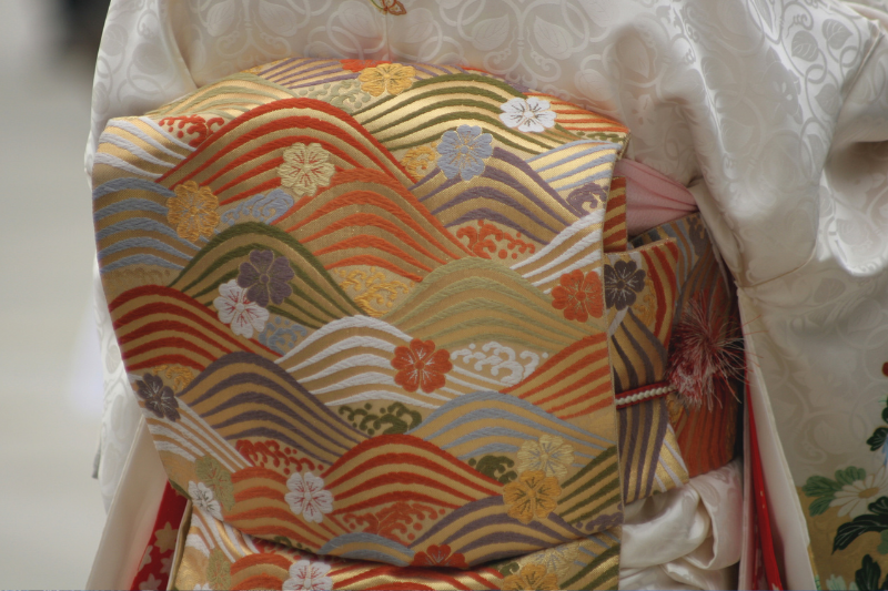 Close-up of a colorful obi with red, gold, green, and purple woven motifs, worn over a white kimono.